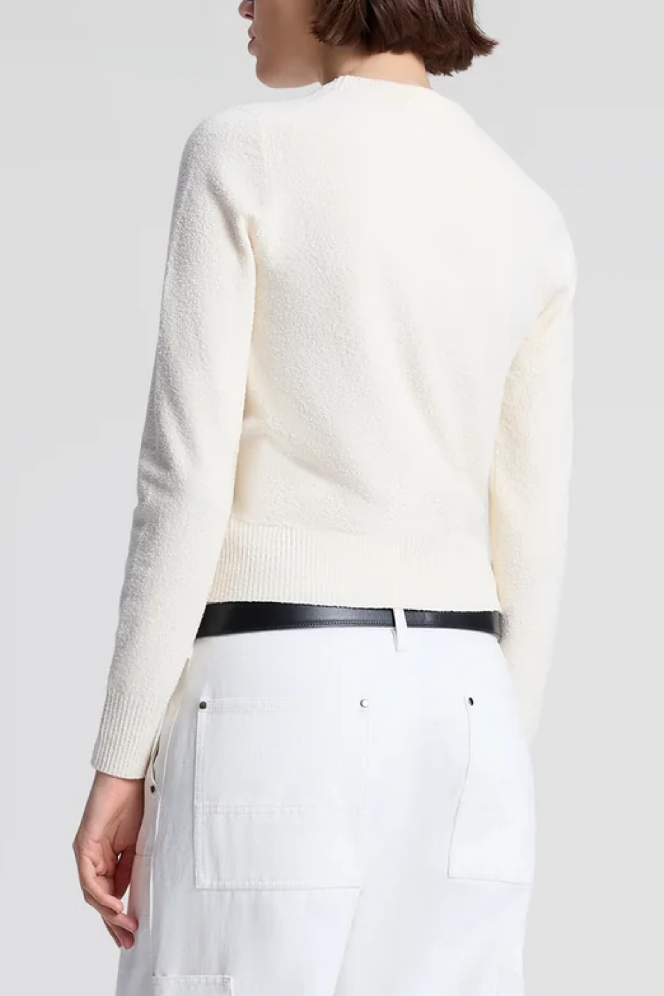 Person wearing a off white sweater and white pants with a black belt on a plain background