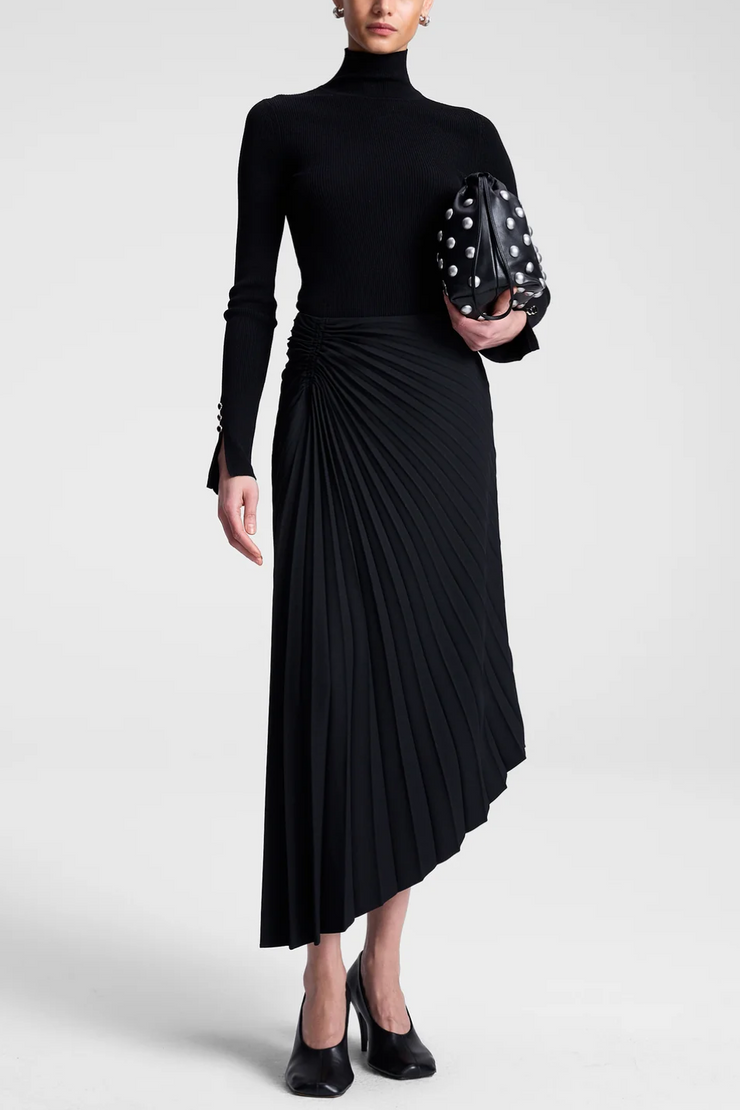 Person wearing a black turtleneck and black pleated skirt holding a patterned clutch.