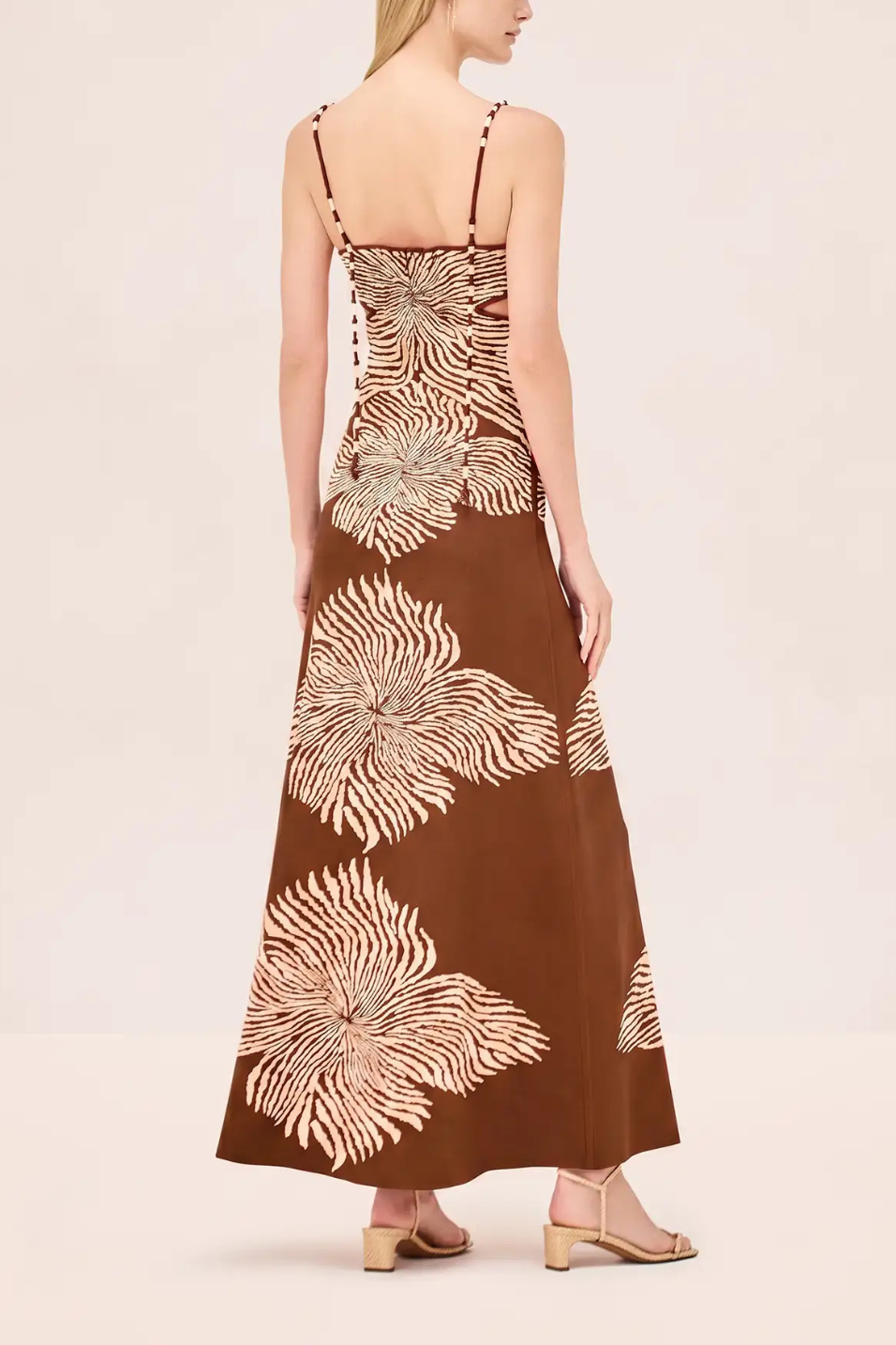 A woman shown from the back wearing a brown maxi dress with a large cream palm leaf pattern, paired with beige heeled sandals, against a solid light beige background.