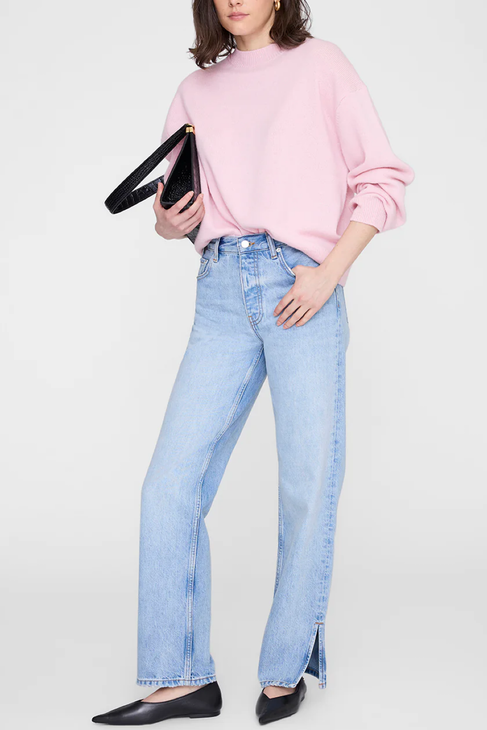 Person wearing a pink sweater and light blue jeans holding a black clutch against a white background