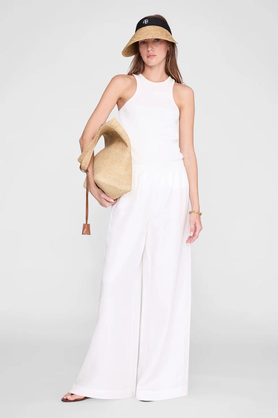 Woman in a visor, white top, and pants holding a beige bag against a white background