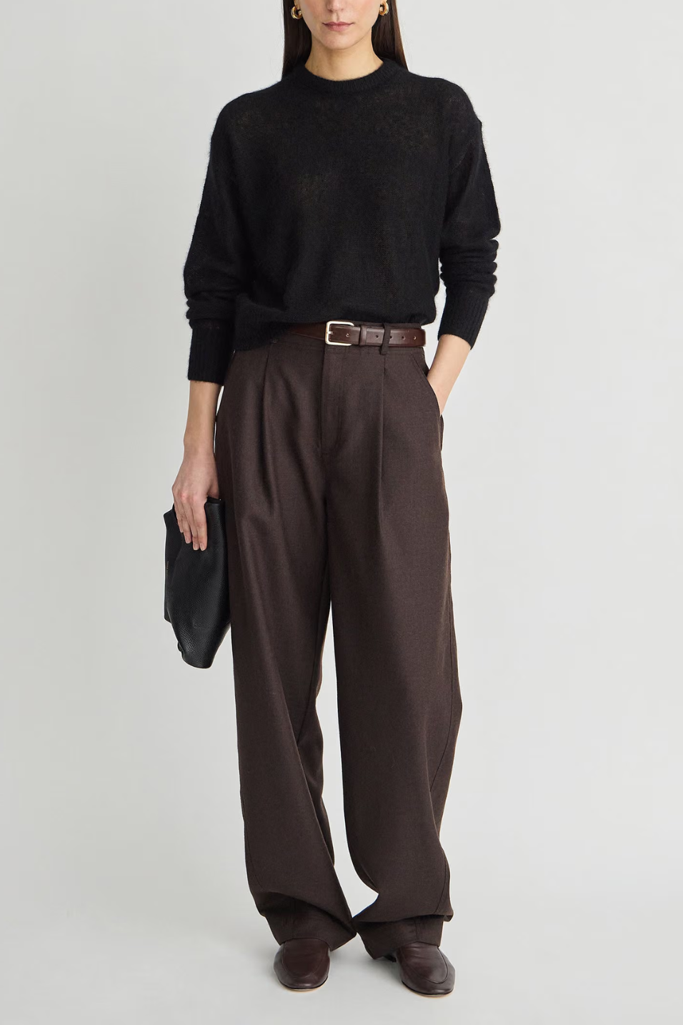 A model wearing a black knit mock-neck sweater, dark brown wide-leg pleated trousers with a brown belt, dark brown leather shoes, and holding a black clutch against a light gray studio background.