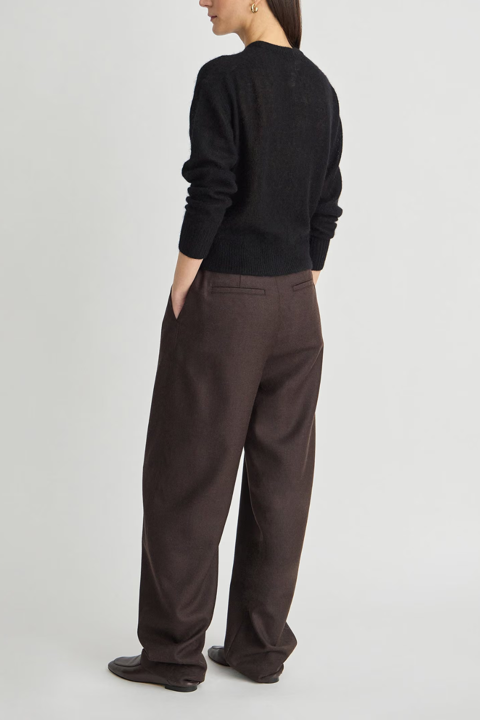 A model shown from the back wearing a black long-sleeve knit sweater and dark brown wide-leg trousers, standing against a light gray studio background.