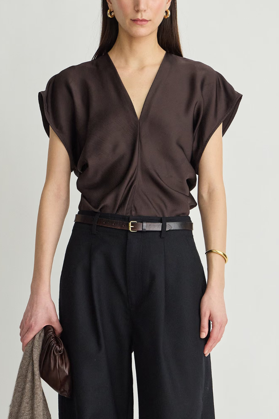 A woman wearing a dark brown wrap-style blouse with short flutter sleeves and black wide-leg trousers, styled with a black belt and holding a brown clutch against a light gray studio background.