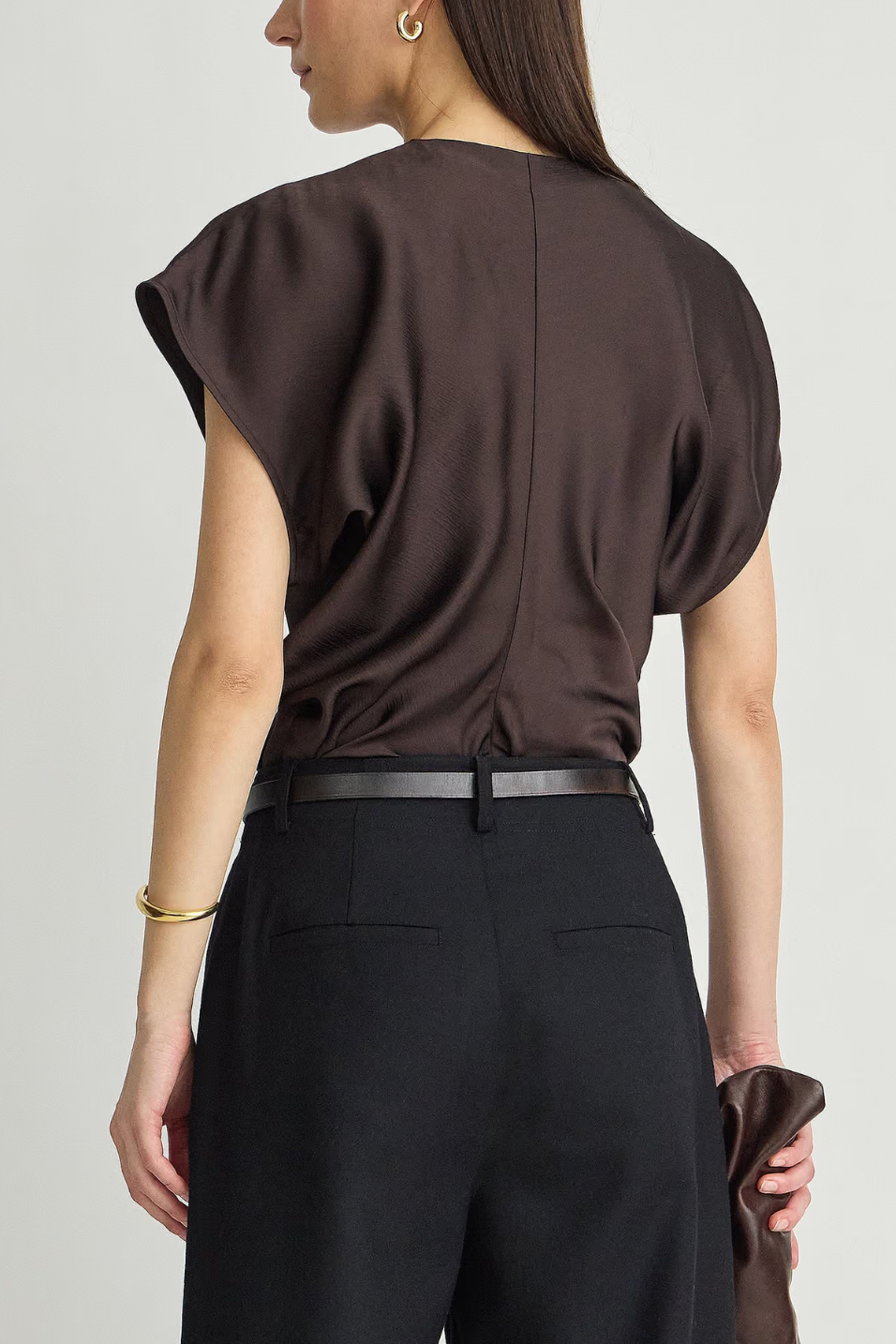 A woman shown from the back wearing a dark brown draped short-sleeve top with a thin black belt and black trousers, against a light gray studio background.