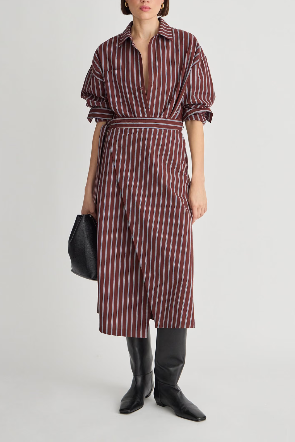 A model wearing a long-sleeved wrap-style midi dress with vertical burgundy and white stripes, styled with black knee-high boots and a black handbag against a light gray studio background.