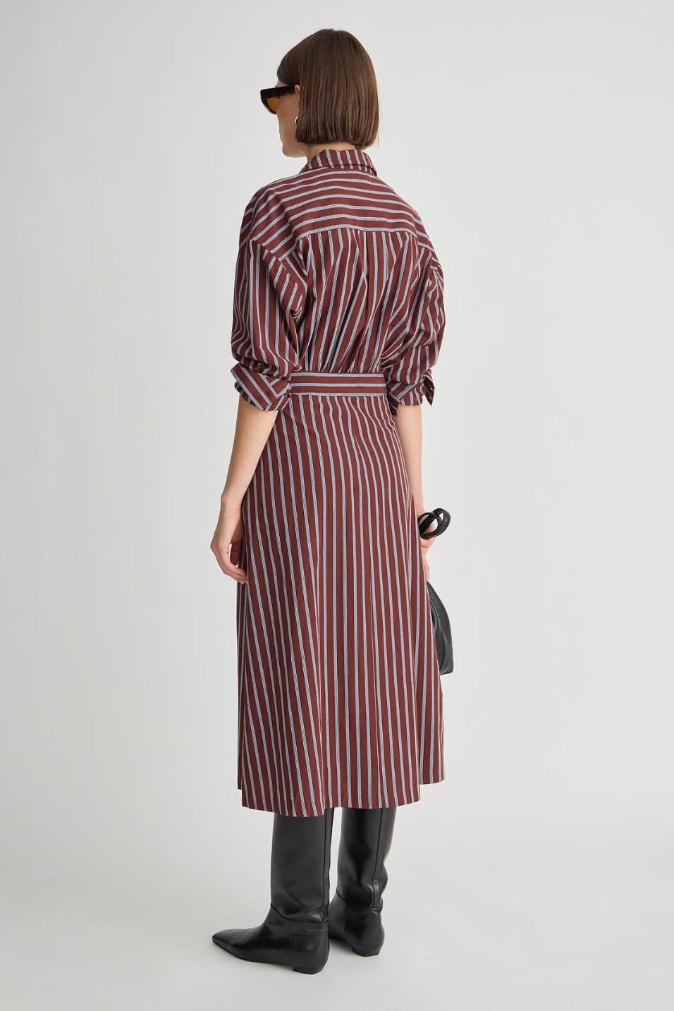 A model shown from the back wearing a long-sleeved midi shirt dress with vertical burgundy, red, and white stripes, styled with black boots and holding a small black handbag against a light gray background.