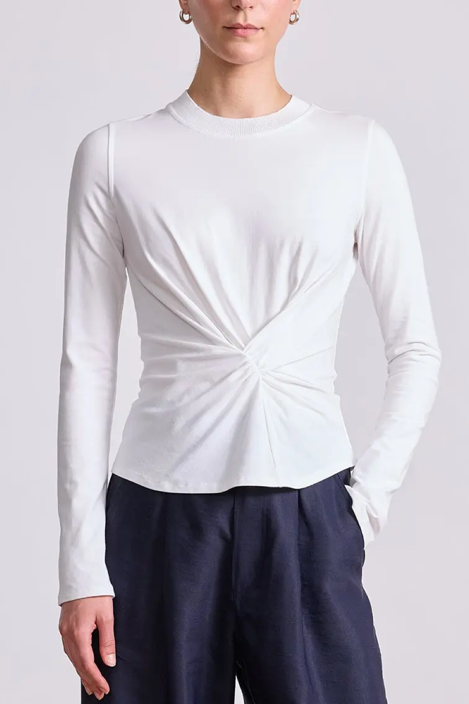 Person wearing a white long-sleeve top with a knot detail and dark pants on a light gray background