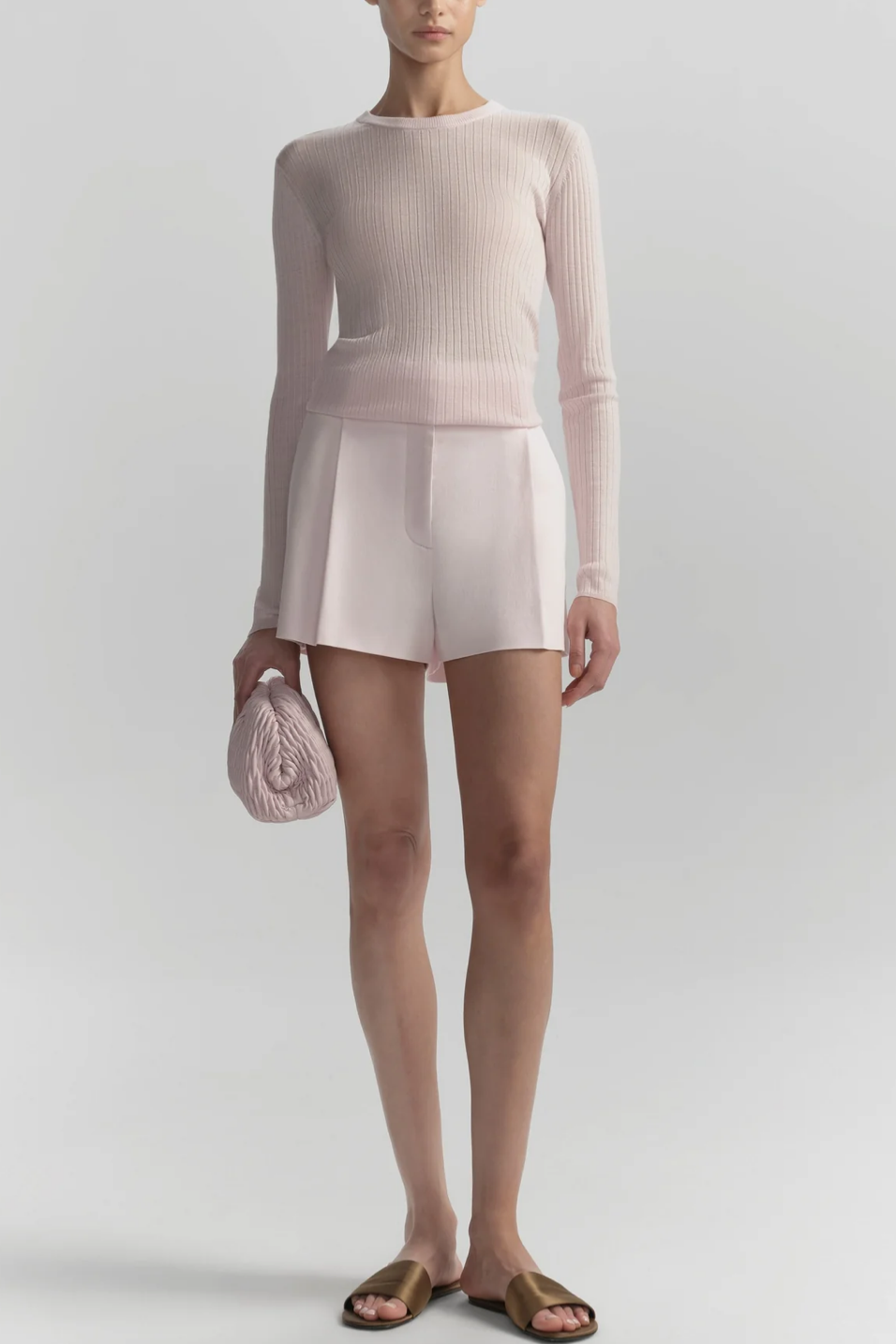 Person wearing a light pink sweater and shorts with a matching clutch against a white background