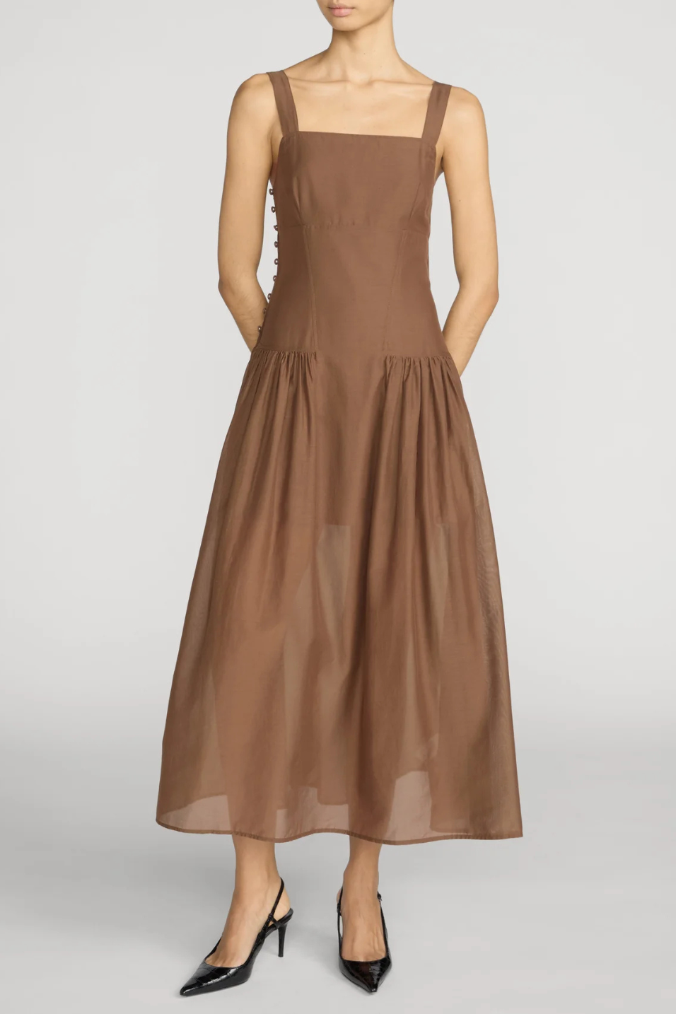 A woman modeling a sleeveless brown midi dress with a square neckline, wide shoulder straps, side buttons, and a flowing skirt, paired with black pointed-toe heels against a light gray studio background.