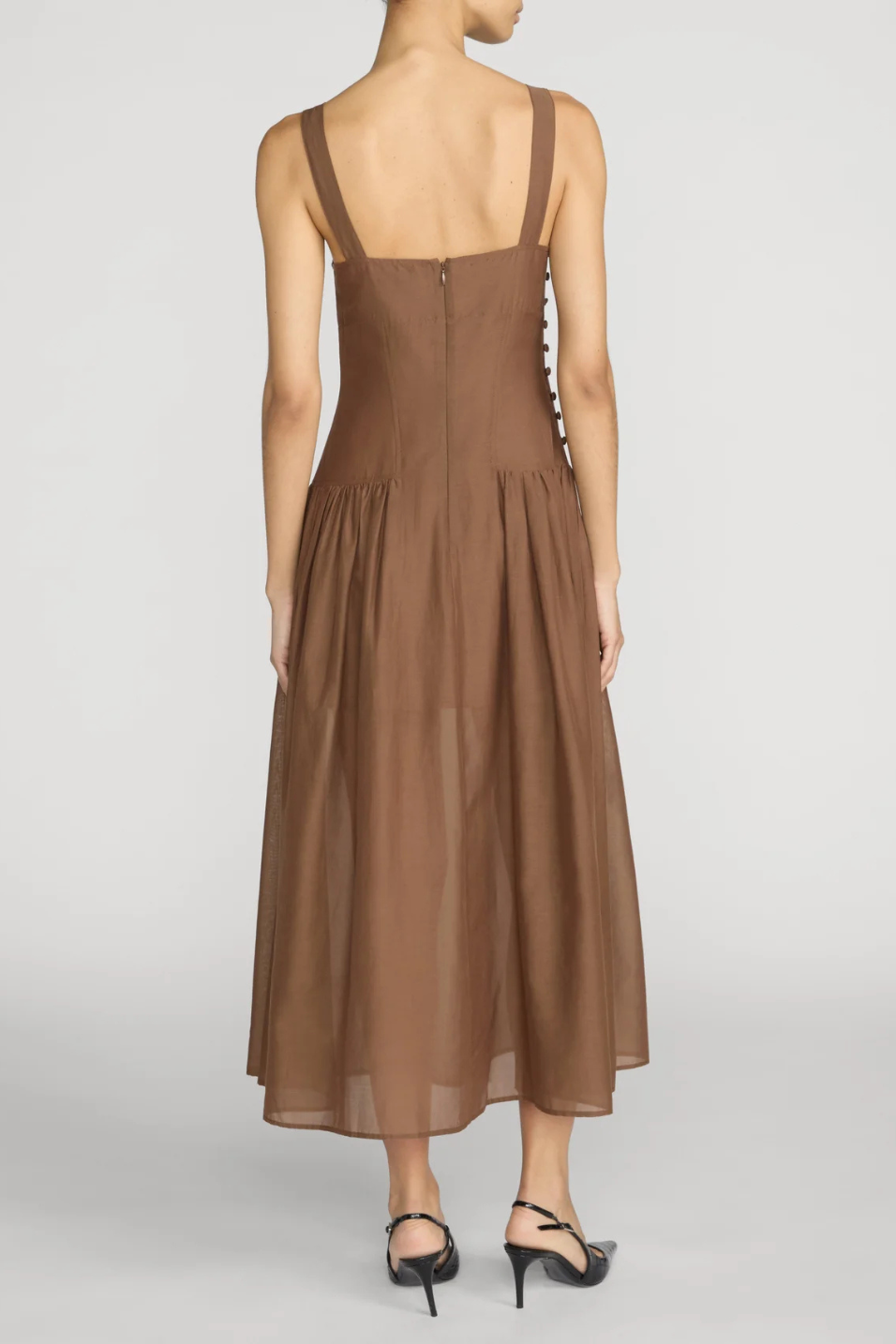 A woman shown from the back wearing a sleeveless brown midi dress with wide shoulder straps, a square neckline, and a gathered skirt. She is standing against a light gray studio background and wearing black high-heeled sandals.