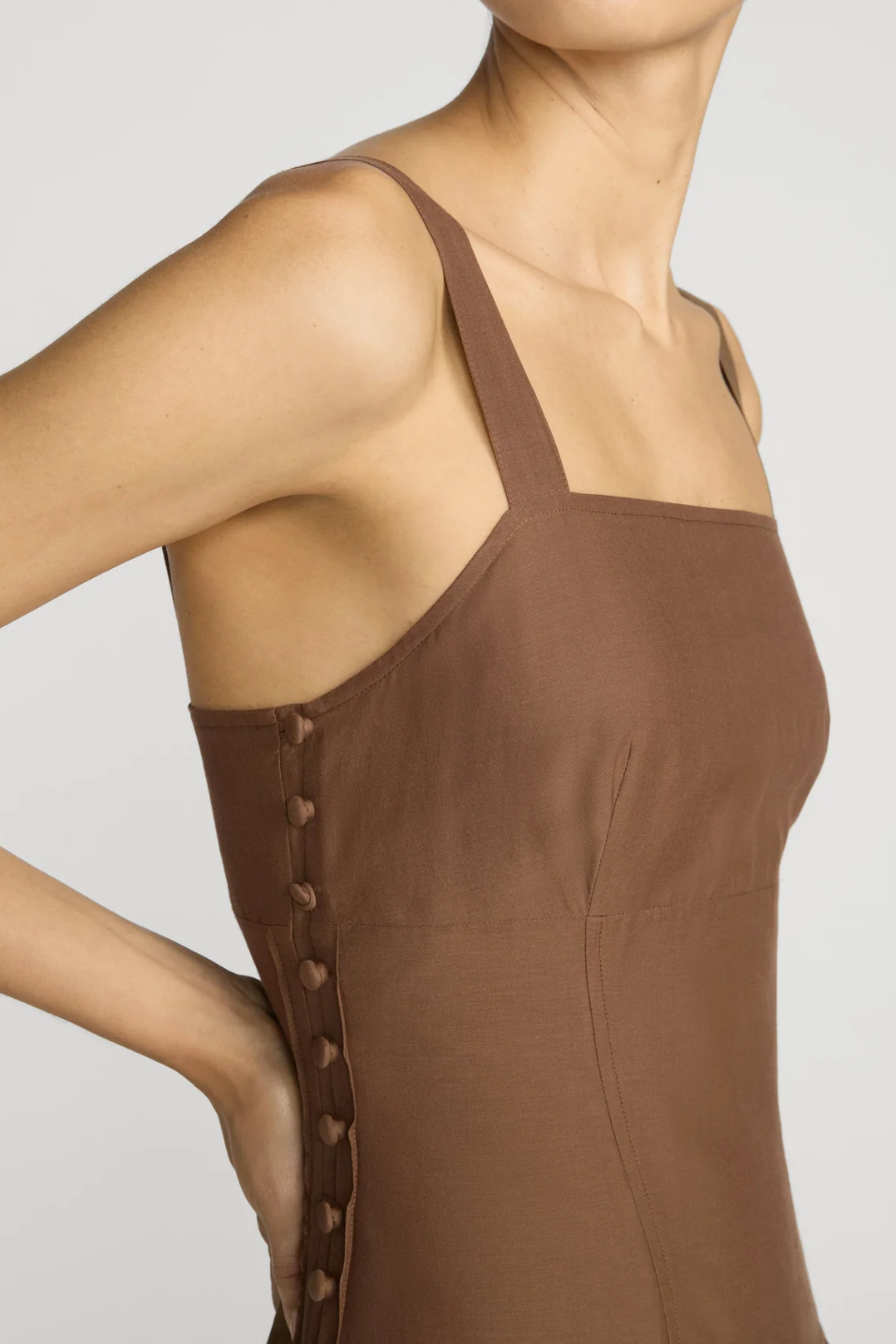 A close-up of a model wearing a brown sleeveless top with square neckline and side buttons, against a light gray background.