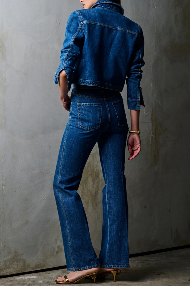 Person wearing a blue denim jacket and jeans against a gray wall.
