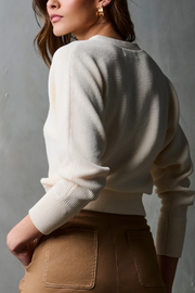 Woman wearing a cream sweater and brown pants against a gray background