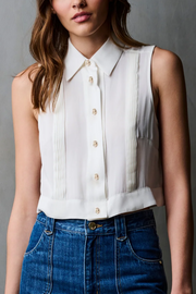 Woman wearing a white sleeveless blouse with gold buttons and blue jeans against a gray background