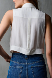 Person wearing a white sleeveless blouse with pleated details and blue jeans against a gray background