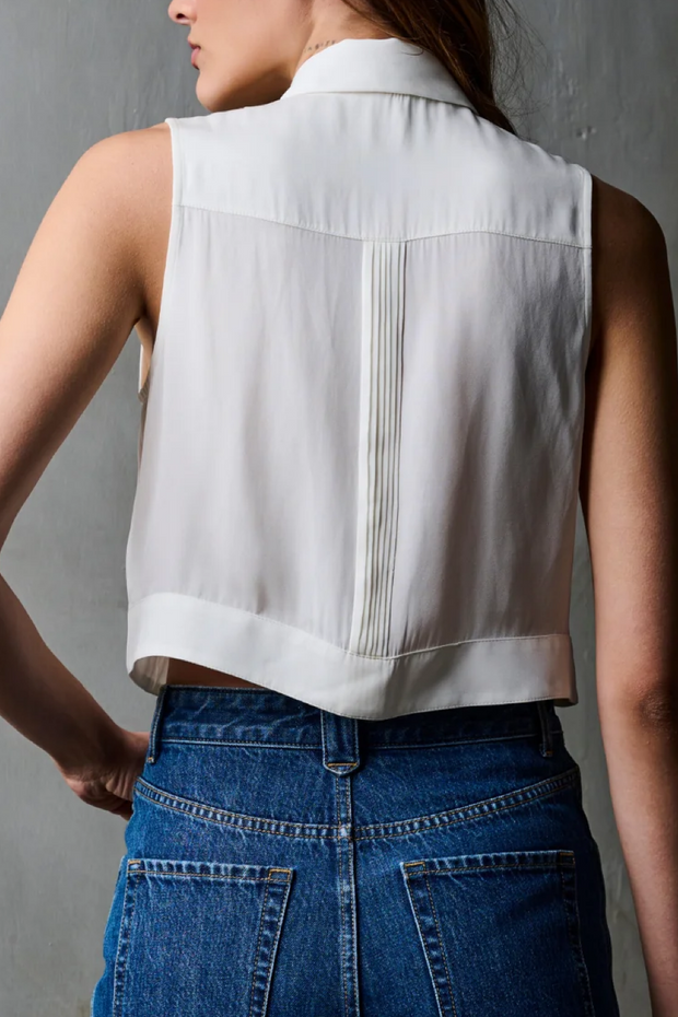 Person wearing a white sleeveless blouse with pleated details and blue jeans against a gray background