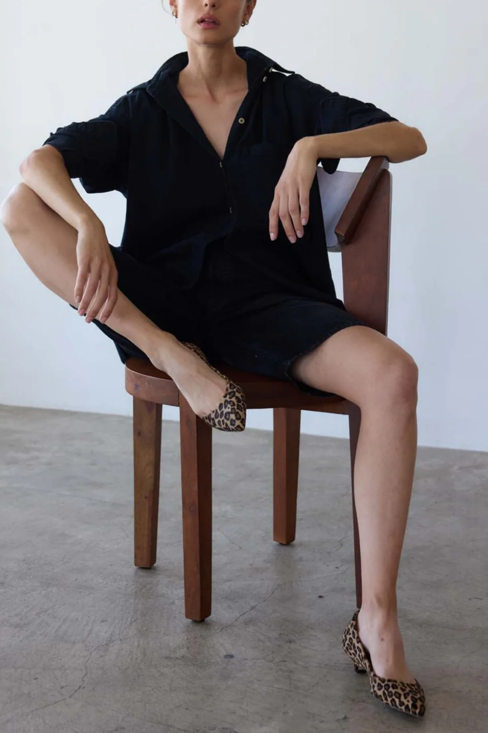 Person wearing a black dress sitting on a wooden chair with leopard print shoes.