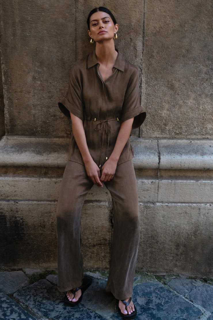 Woman in a mocha top and pants sitting against a stone wall.