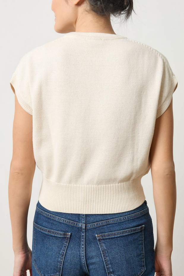 Person wearing a cream sleeveless sweater and blue jeans on a plain background