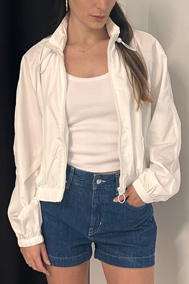 Person wearing a white jacket, white top, and blue shorts against a neutral background
