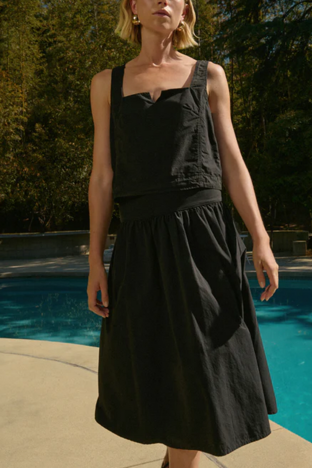 Woman wearing a black tank and skirt standing by a pool with trees in the background