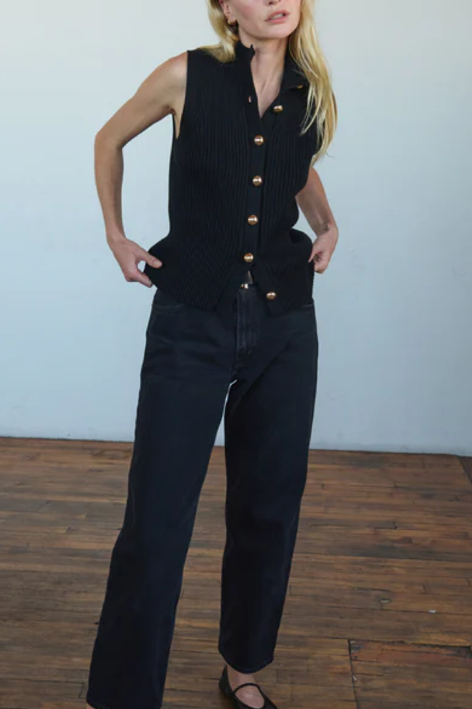 Person wearing a black sleeveless vest with gold buttons and dark jeans on a wooden floor.