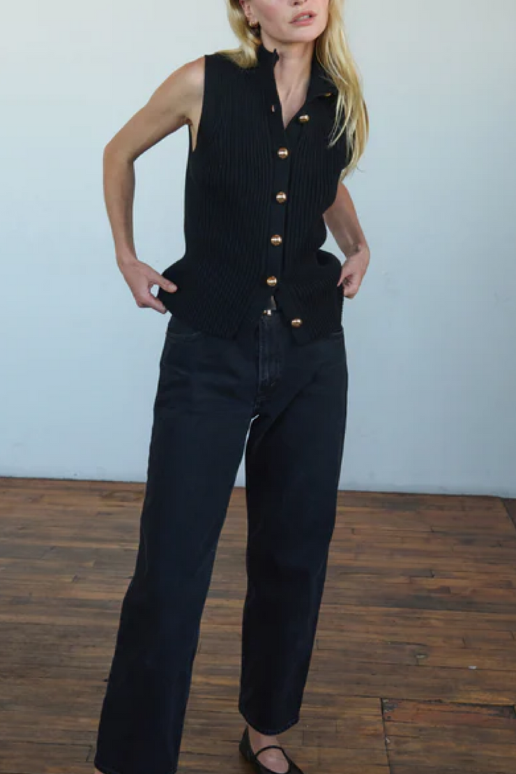 Person wearing a black sleeveless vest with gold buttons and dark jeans on a wooden floor.