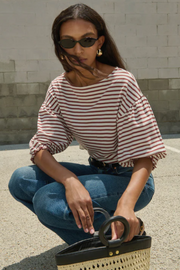 Woman in a striped shirt and jeans squatting with a woven handbag against a stone wall.