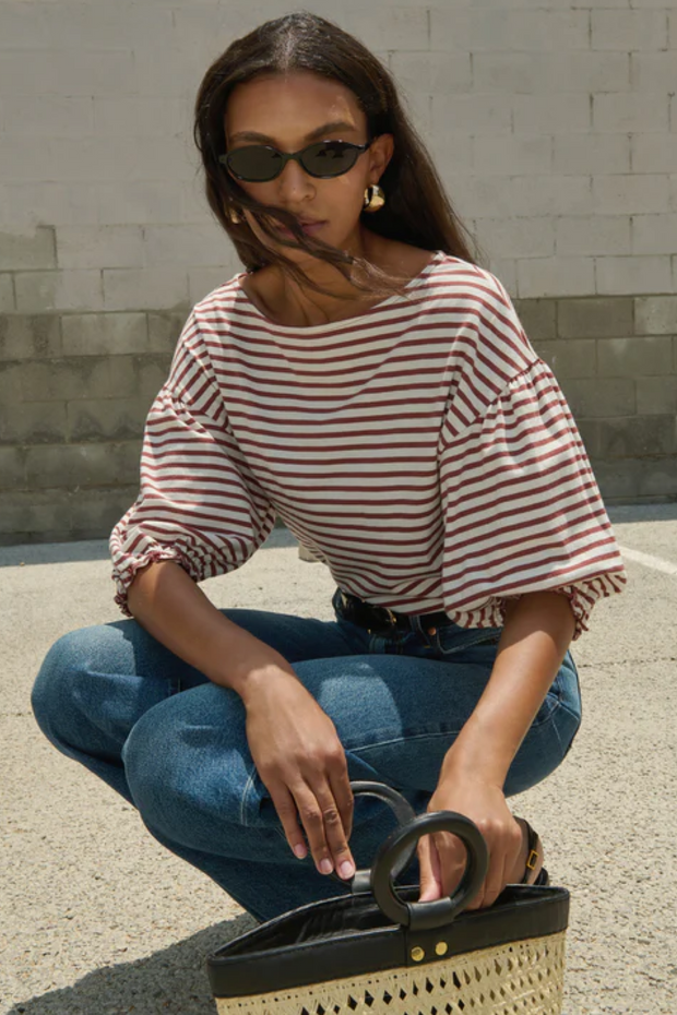 Woman in a striped shirt and jeans squatting with a woven handbag against a stone wall.
