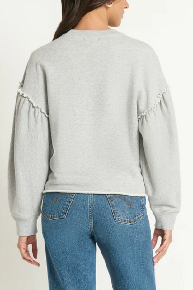 Person wearing a light gray sweatshirt with ruffled sleeves and blue jeans on a white background