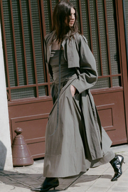 Woman wearing a long, light-colored coat and khaki dress standing in front of a building with a brown door.