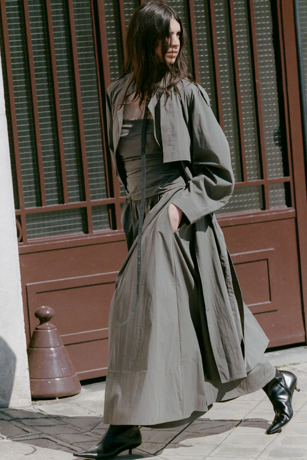 Woman wearing a long, light-colored coat and khaki dress standing in front of a building with a brown door.