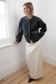 Woman wearing a black jacket and white skirt standing in a room with wooden flooring and a window.