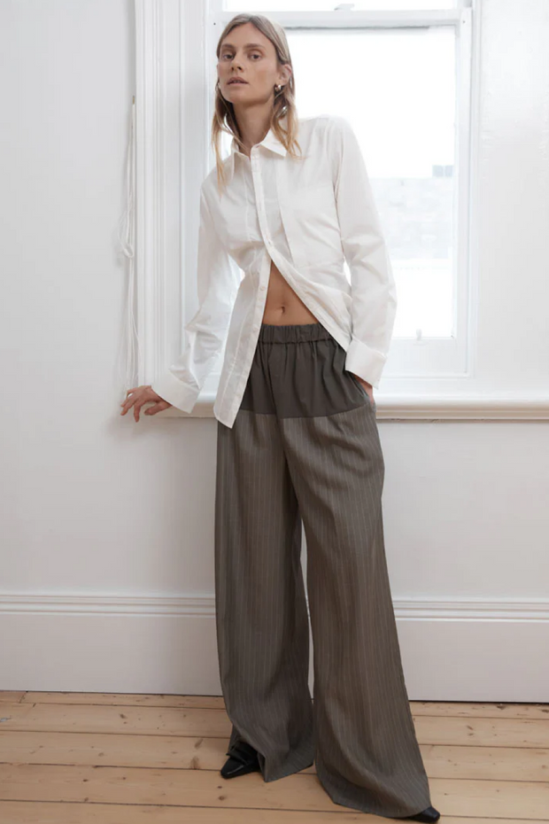 Person wearing a white shirt and khaki pinstripe wide-leg pants standing in a room with a window.