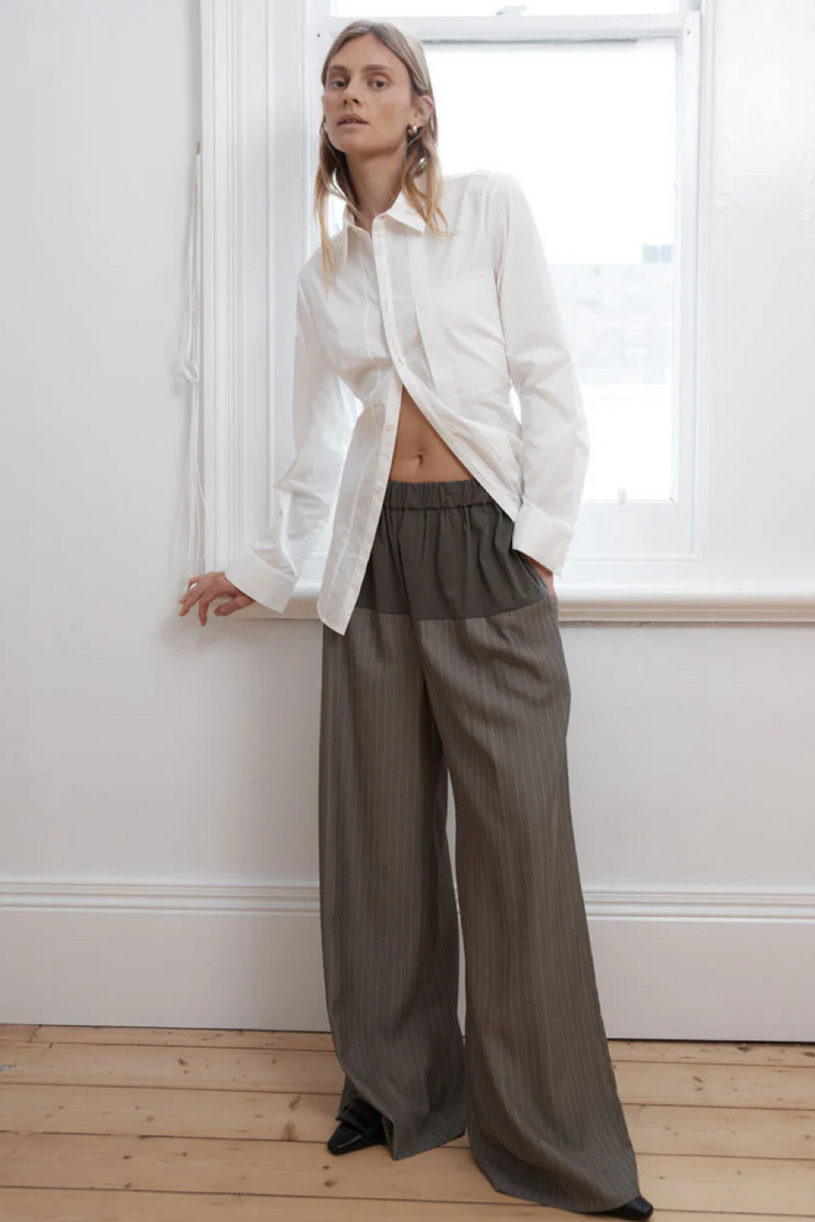 Person wearing a white shirt and khaki pinstripe wide-leg pants standing in a room with a window.