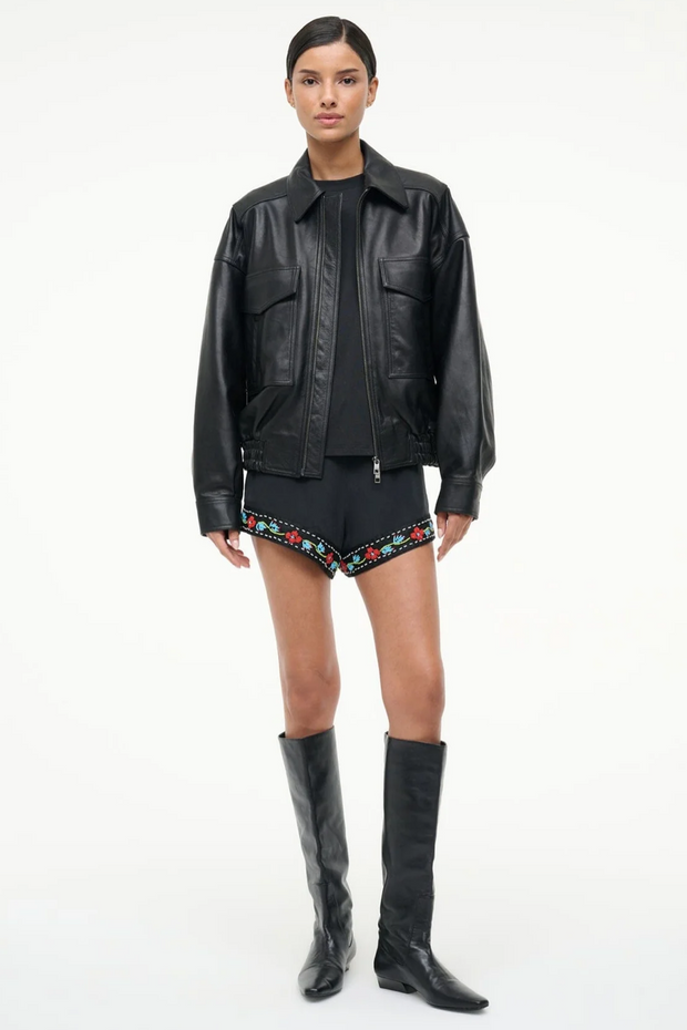 Person wearing a black leather jacket, black top, black shorts with floral embroidery, and black knee-high boots on a white background.