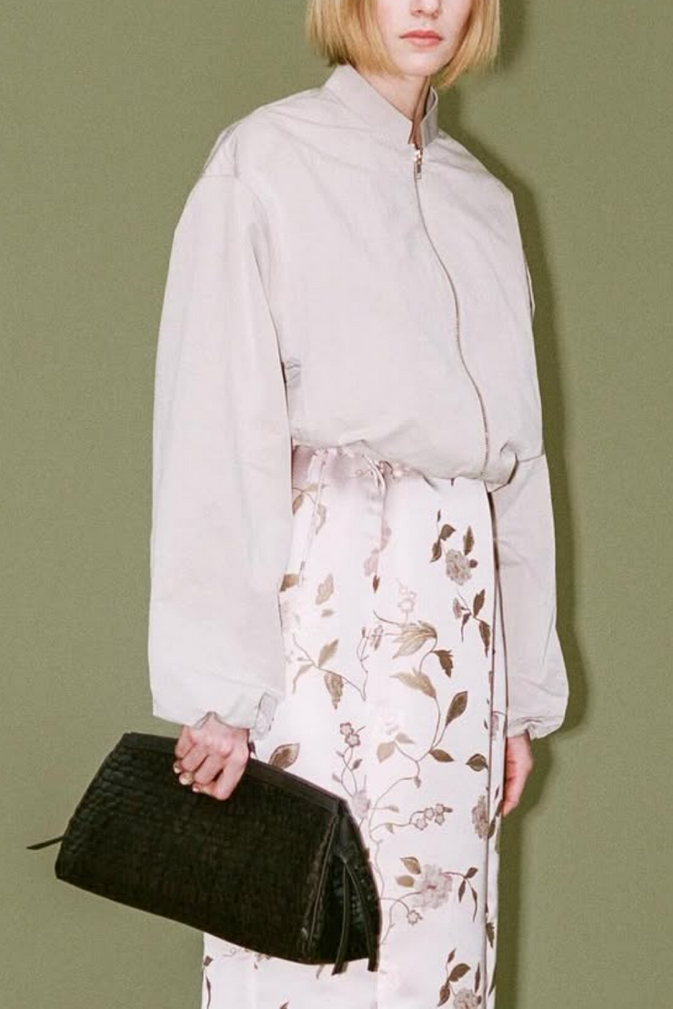 Person wearing a jacket over a  floral dress and holding a clutch against a green background