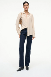 Woman wearing a beige shirt, dark jeans, and black shoes on a white background