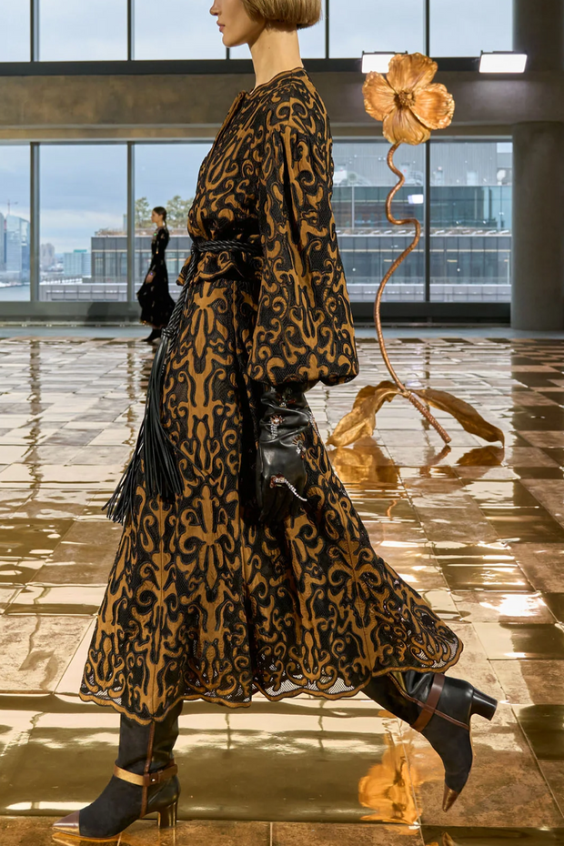 Woman in a printed jacket and skirt standing in a modern indoor setting with large windows.