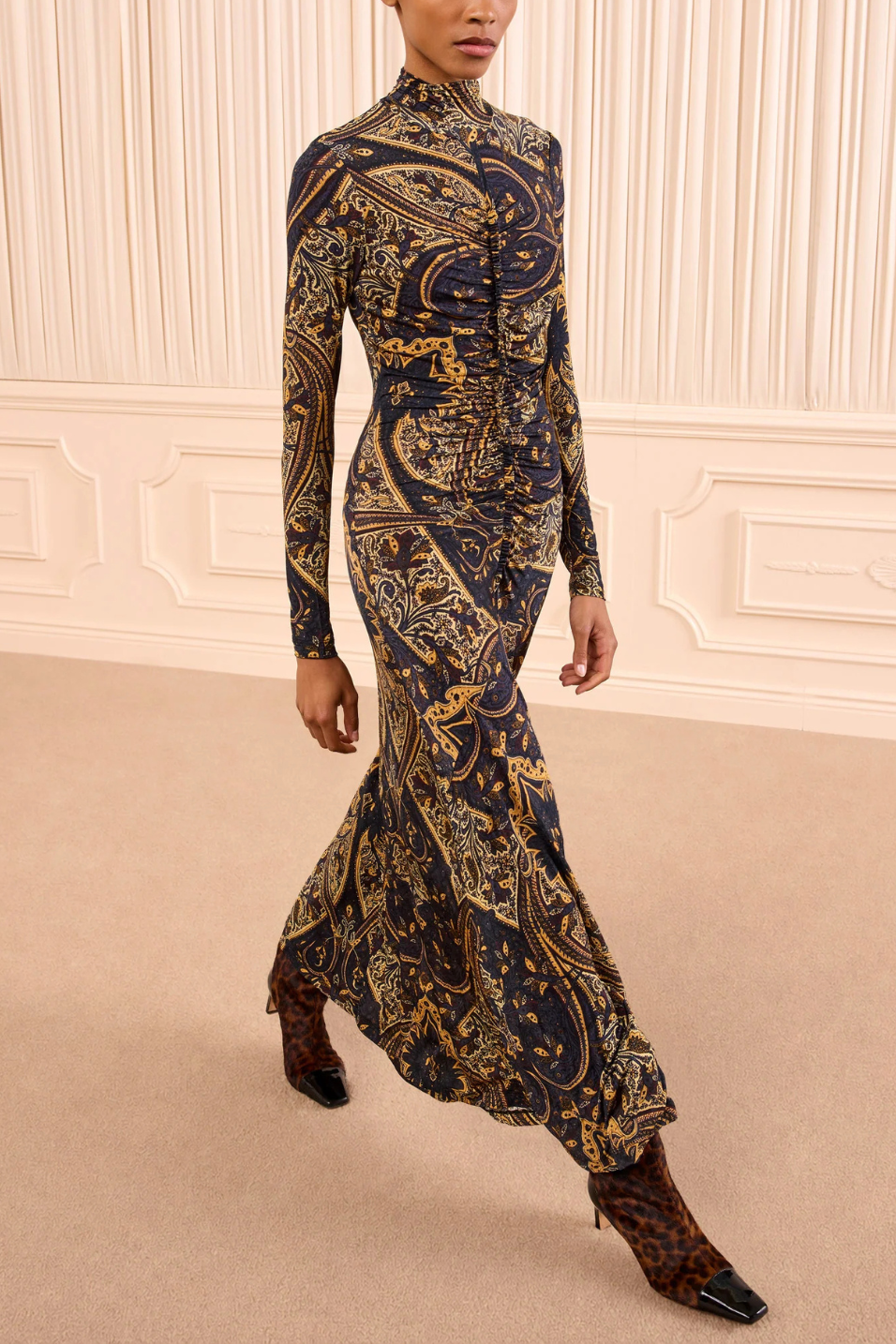 A model wearing a long-sleeved, high-neck maxi dress with an intricate gold and brown paisley pattern on a black base, paired with brown pointed-toe heels, set against a cream-colored paneled wall.