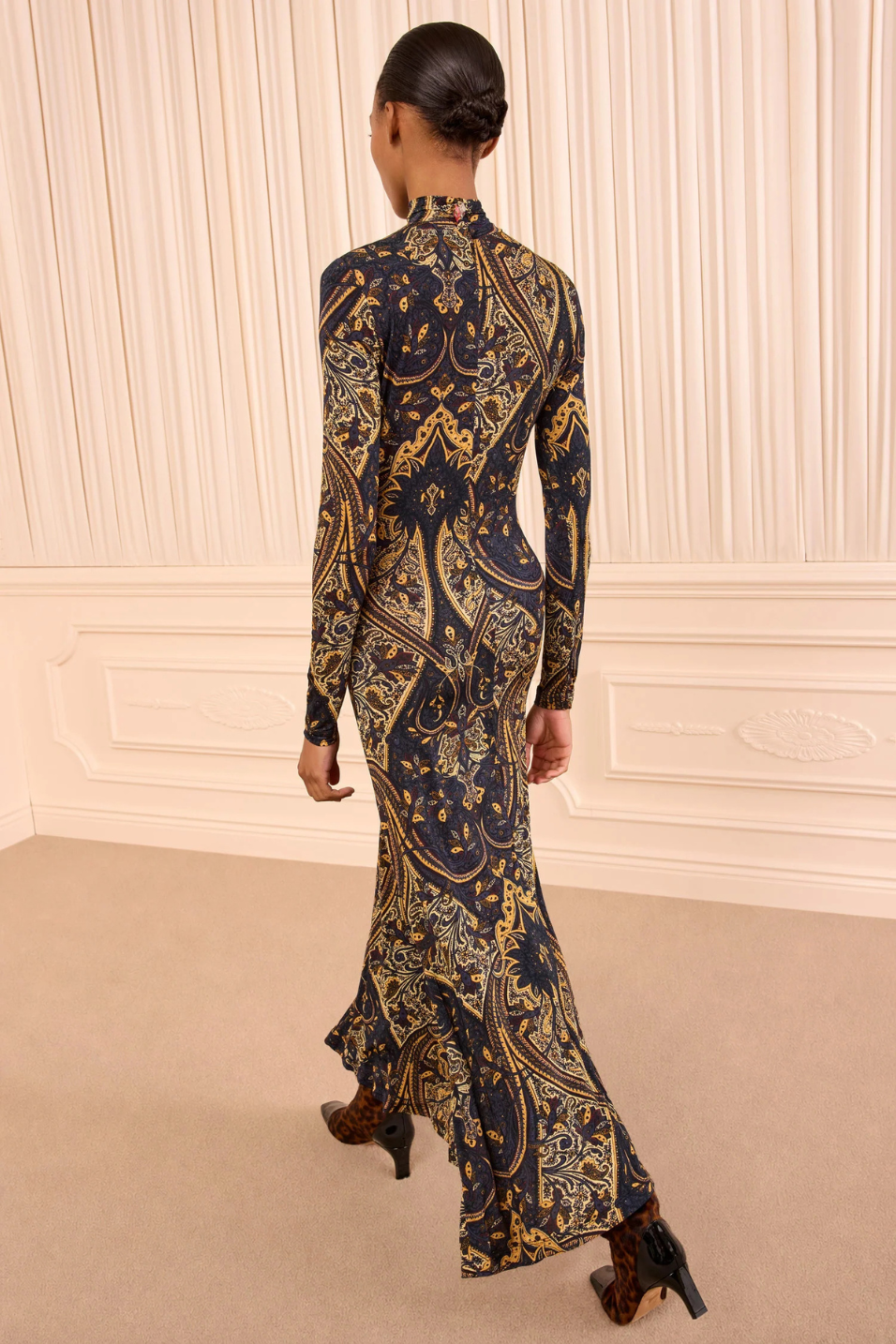 A woman viewed from behind wearing a long-sleeve high-neck maxi dress with an intricate navy blue, gold, and beige baroque-style pattern, standing in a room with cream paneled walls and beige carpet.