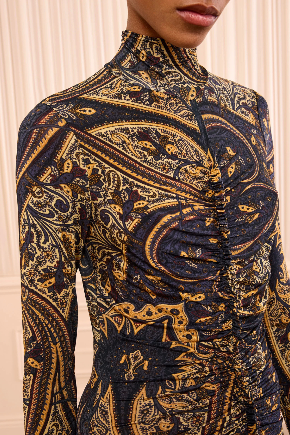 A close-up of a model wearing a long-sleeve mock-neck top with a dark navy blue and gold paisley pattern, featuring ruched detailing on the chest and waist.