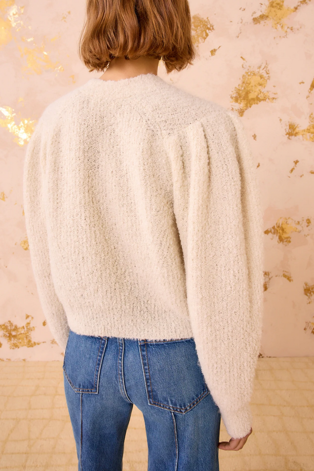 Person wearing a cardigan and blue jeans against a floral-patterned wall.