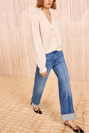 Person wearing a cardigan and blue jeans standing against a textured beige wall.