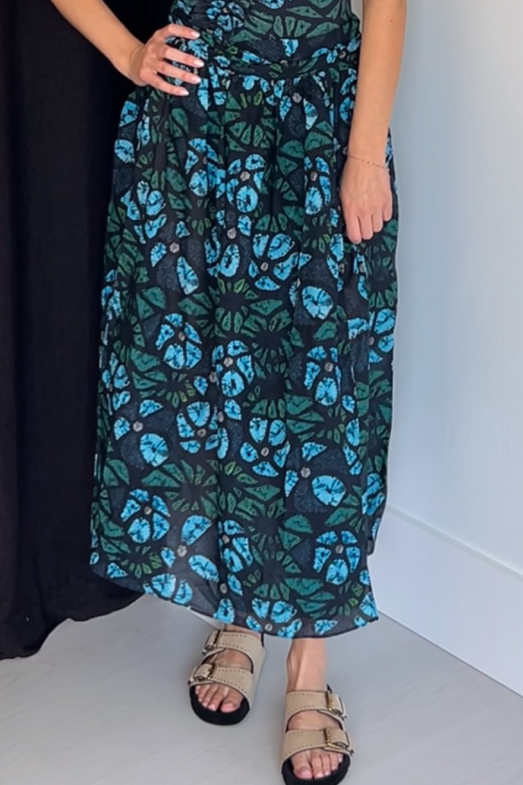 Person wearing a long floral cover-up  sarong-skirt with blue and green patterns on a plain background