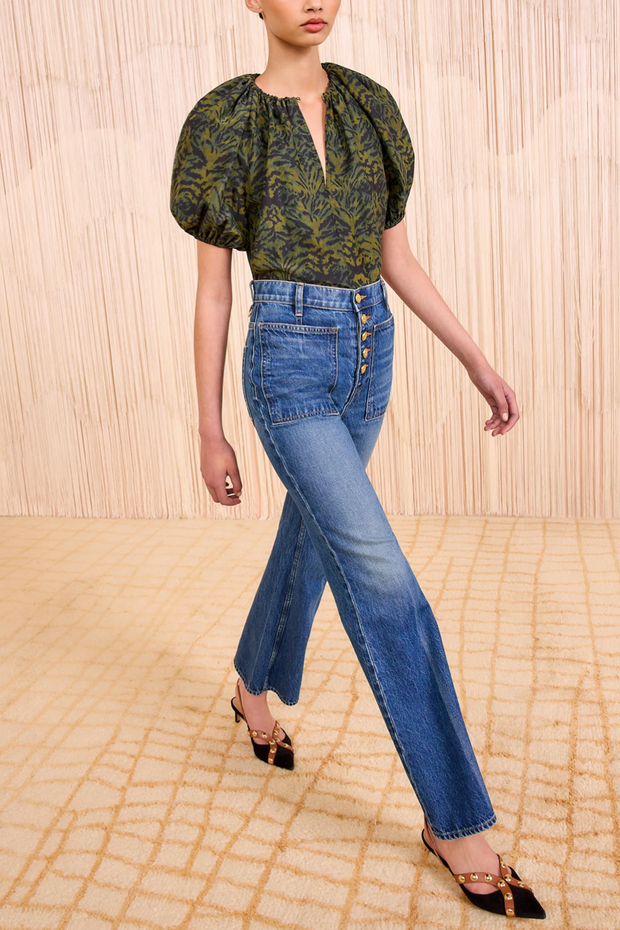 Woman wearing a green patterned top and blue jeans against a beige wall.