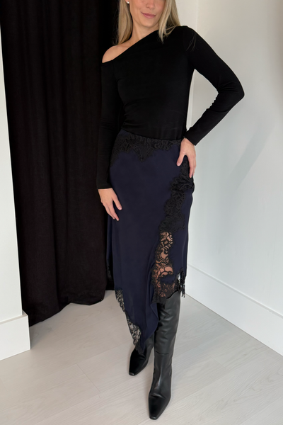 Woman wearing a black off-shoulder top and navy skirt with lace details, standing against a plain background.