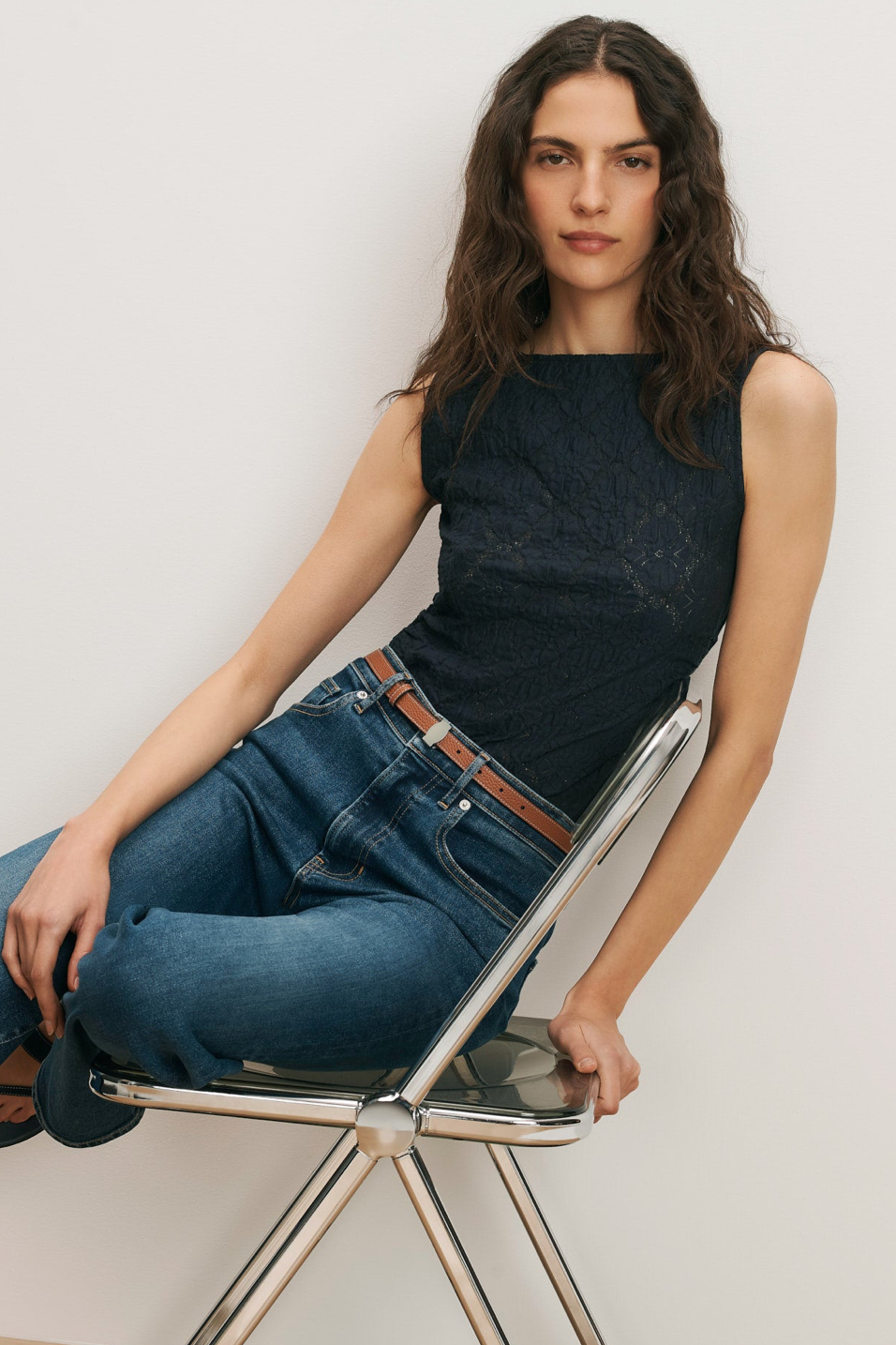 Woman sitting on a chair wearing a black sleeveless top and blue jeans against a plain background