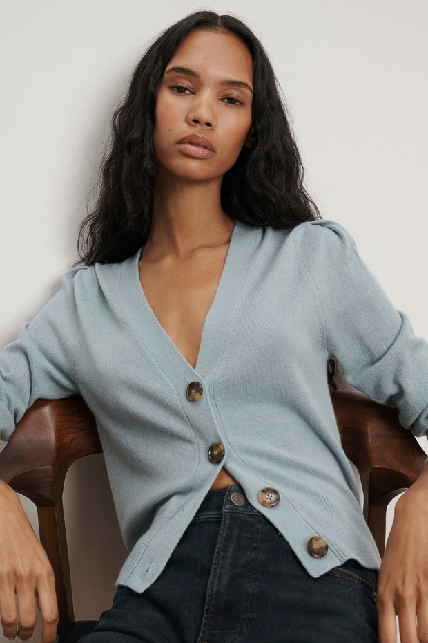 Woman wearing a light blue cardigan with buttons, sitting on a brown chair against a plain background.